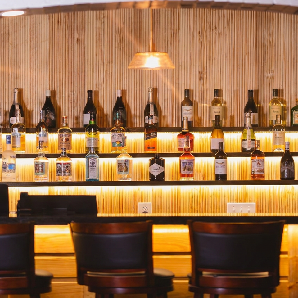 Warmly lit restaurant bar displaying rows of liquor bottles on illuminated wooden shelves, with dark bar stools in front.